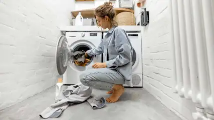 Een vrouw hurkt bij een open wasmachine in een witte kamer met een witte radiator.
