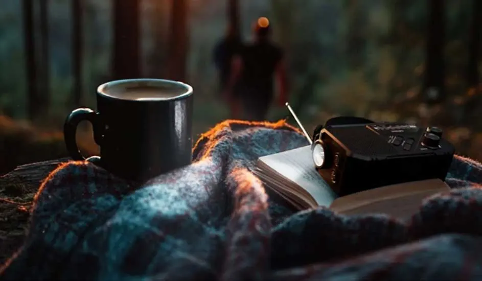 Op een plaid in het bos staan een dampende mok, een geopend notitieboek en een compacte radio, badend in zacht avondlicht.
