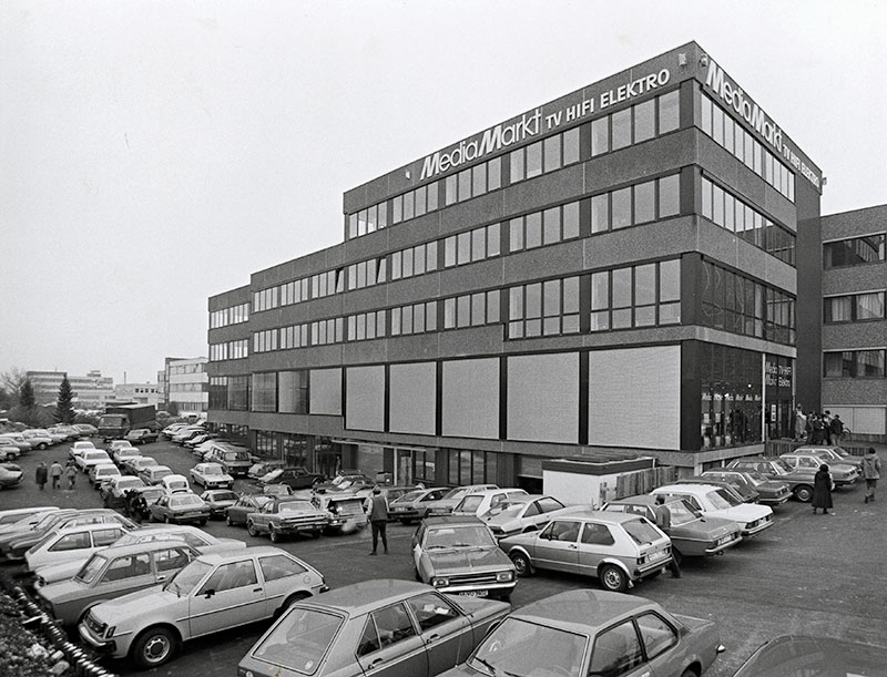 Grijstinten foto van een Media Markt gebouw met auto's op de parkeerplaats en mensen die rondlopen.