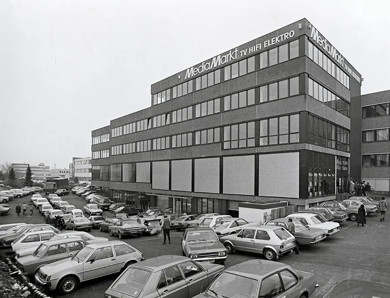Grijstinten foto van een Media Markt gebouw met auto's op de parkeerplaats en mensen die rondlopen.