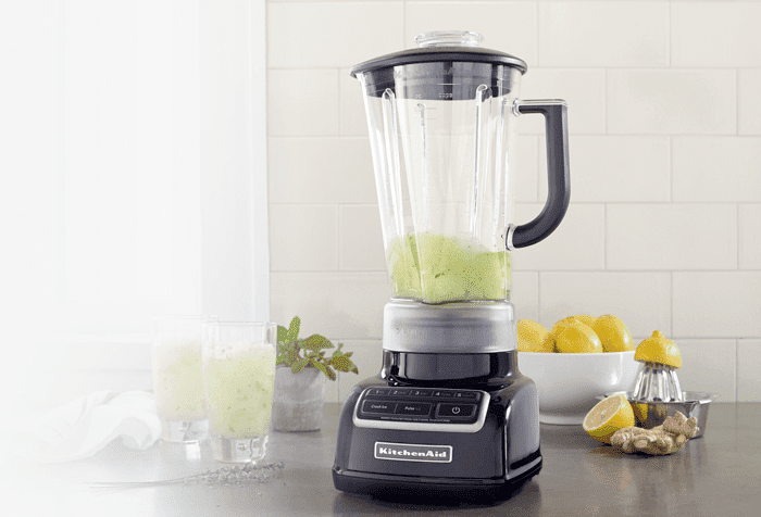 Een zwarte KitchenAid blender met een kan gevuld met een groene vloeistof staat op een aanrecht. Er staan citroenen en gember naast.