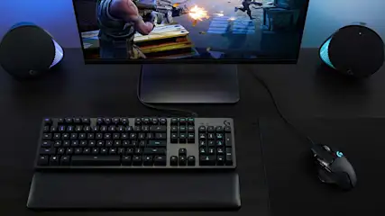 Donkere gaming opstelling met groot scherm, RGB toetsenbord, muis en krachtige luidsprekers, terwijl een actiespel vol explosies wordt gespeeld.