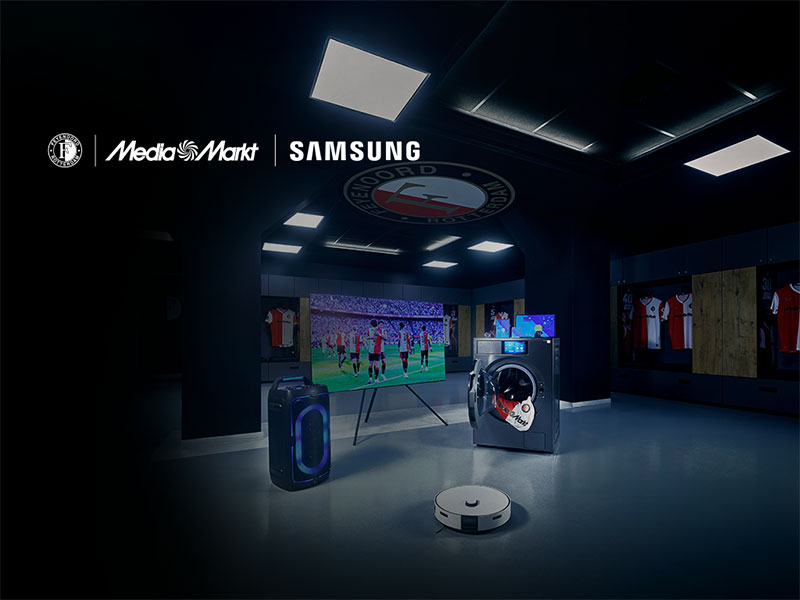 Donkere clubwinkel- of kleedkamerachtige ruimte met MediaMarkt- en Samsung-logo’s, een tv waarop een voetbalwedstrijd te zien is, en Samsung-apparaten op de vloer (speaker, wasmachine en robotstofzuiger); op de achtergrond hangen rood-witte voetbalshirts.
