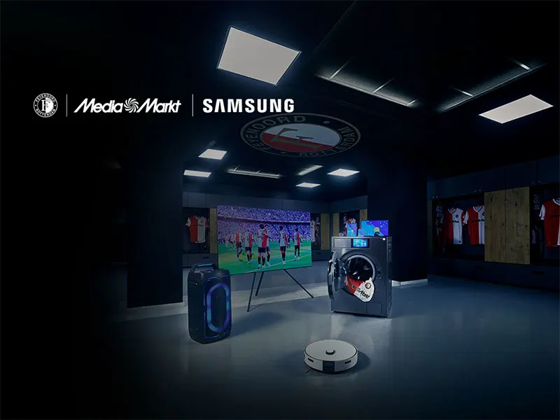 Donkere clubwinkel- of kleedkamerachtige ruimte met MediaMarkt- en Samsung-logo’s, een tv waarop een voetbalwedstrijd te zien is, en Samsung-apparaten op de vloer (speaker, wasmachine en robotstofzuiger); op de achtergrond hangen rood-witte voetbalshirts.