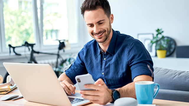 Een man zit aan een bureau met een laptop en een telefoon, en een blauwe mok staat ernaast. Achter hem staat een fiets.
