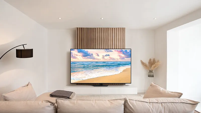 Een woonkamer met een tv waarop een strand te zien is, een vloerlamp en een beige bank.