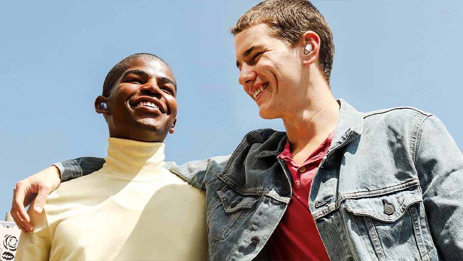 Twee mannen lachen, beiden met oortjes in. Een draagt een beige coltrui, de ander een rood shirt en een spijkerjack. De lucht is blauw.
