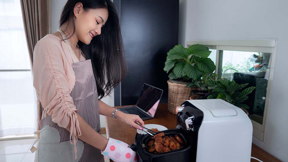 Een vrouw met een schort gebruikt een airfryer in een keuken met een laptop, planten en een raam.
