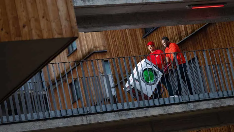 Twee mensen in rode shirts vervoeren een wasmachine op een balkon met een metalen hek. Houten gebouw op de achtergrond.