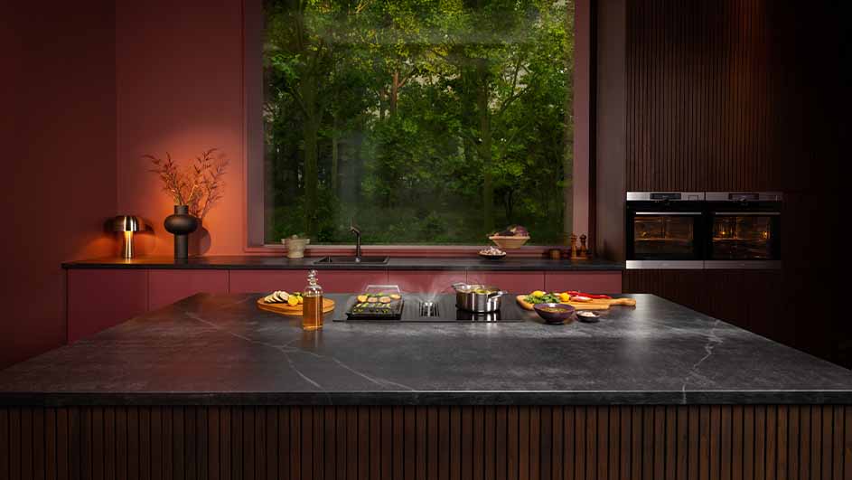 Moderne keuken met kookeiland, kookplaat, groenten en pannen. Een groot raam biedt uitzicht op bomen. Twee ovens zijn zichtbaar.