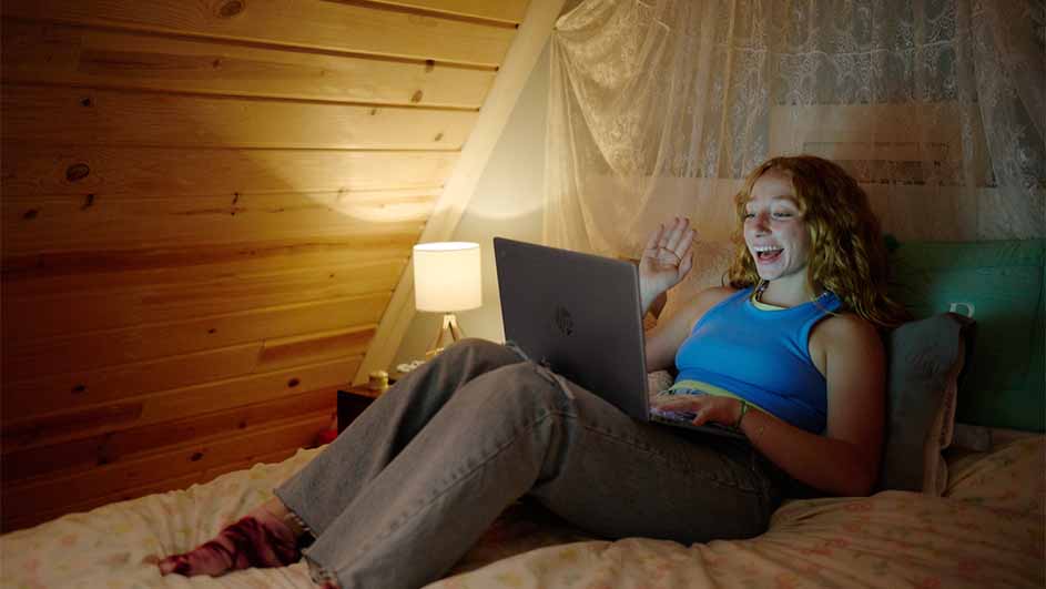 Vrouw zit op een bed met een laptop, zwaait. Houten wand, lamp en gordijnen op de achtergrond.