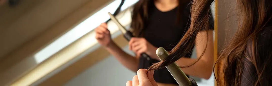 Person styling long brown hair with a curling iron in a well-lit room, wearing black clothing.