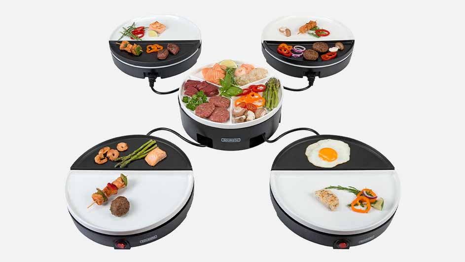 Vier kleine, zwart-witte raclette-grills zijn aangesloten op een centrale schaal met vlees, vis en groenten.