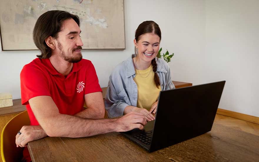 Two people are smiling at a laptop at a wooden table, one in a red shirt and one in a yellow shirt with a denim jacket.
