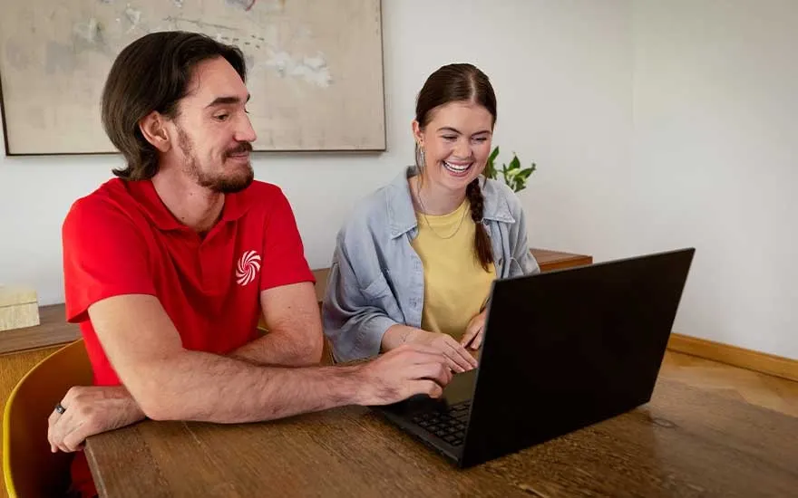 Employee in red polo supports a customer at a wooden table, explaining a laptop while they both share a smile.