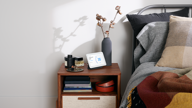 Nachtkastje met boeken, decoratie, een vaas met takken, en een digitaal scherm naast een bed met kussens en een deken.