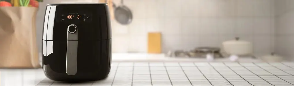 A black air fryer sits on a white tiled countertop. A bag of groceries is to the left. The background shows a kitchen setting.