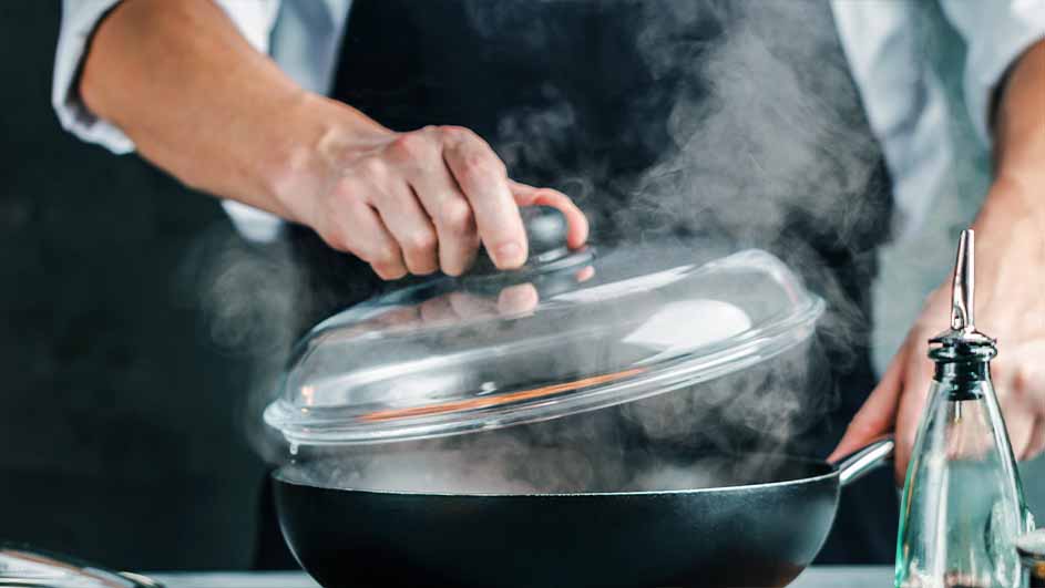 Een persoon met een zwart schort tilt de doorzichtige deksel van een zwarte pan waar stoom uitkomt, naast een glazen fles.