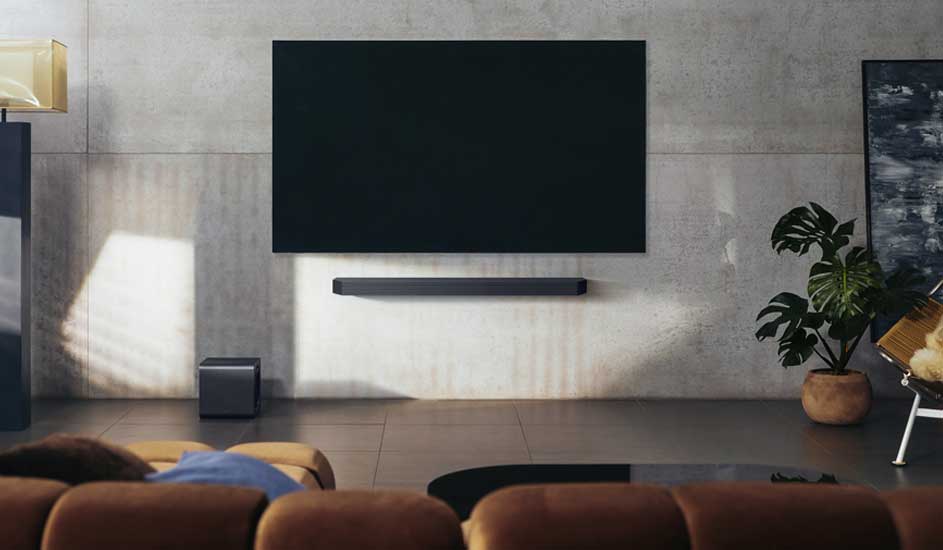 Een moderne woonkamer met een grote zwarte tv aan de muur, een soundbar eronder, een subwoofer links en een plant rechts op de vloer.