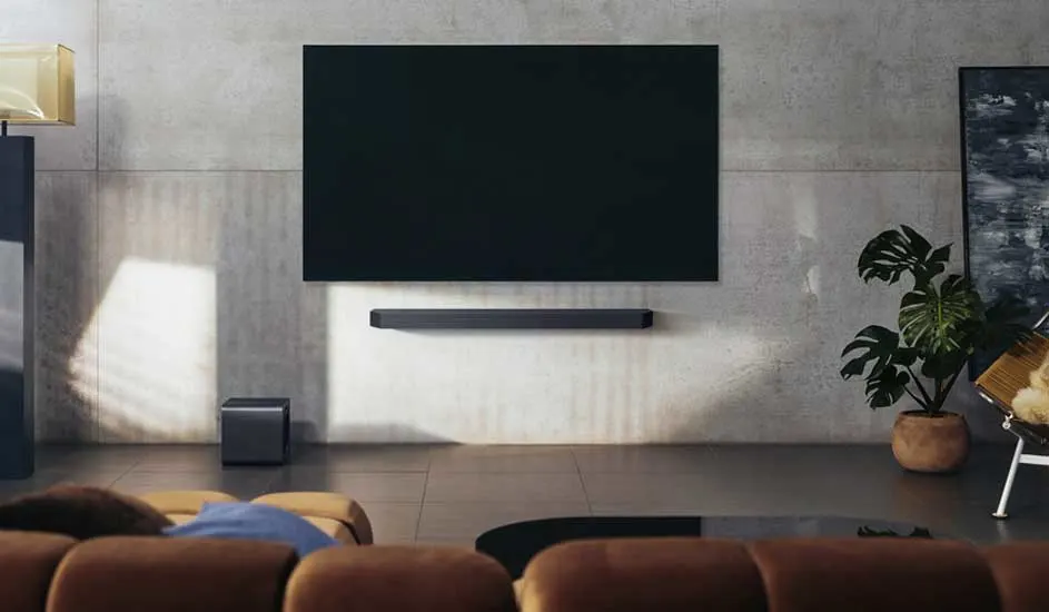 Een moderne woonkamer met een grote zwarte tv aan de muur, een soundbar eronder, een subwoofer links en een plant rechts op de vloer.