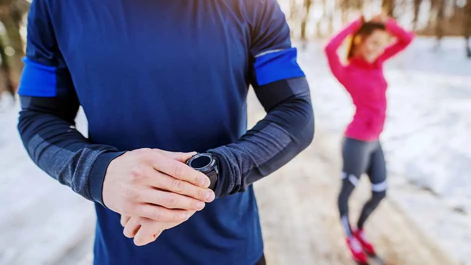 Man controleert horloge in blauw shirt. Vrouw in roze shirt rekt zich uit op de achtergrond in een winterlandschap.