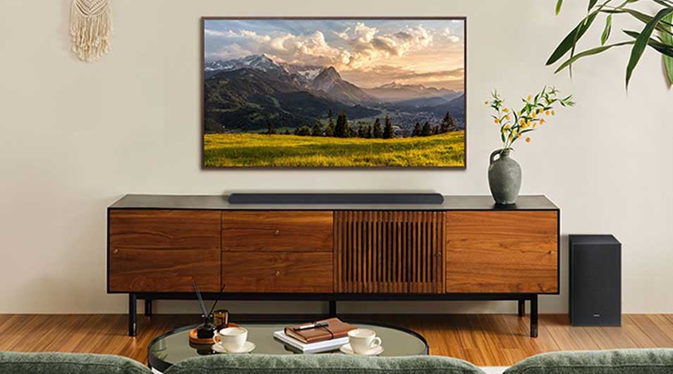 Moderne woonkamer met tv boven houten dressoir, soundbar, subwoofer, planten en berglandschap op het scherm.