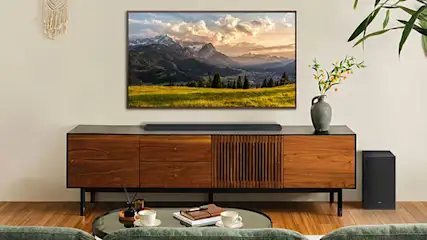 Moderne woonkamer met tv boven houten dressoir, soundbar, subwoofer, planten en berglandschap op het scherm.