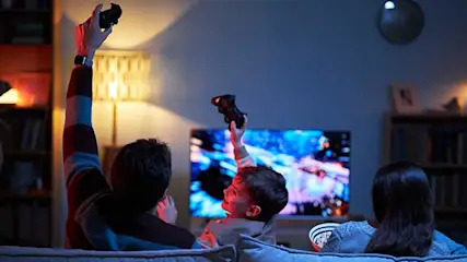 A family sit on the sofa, excitedly gaming or watching a video game on television in a cosy room.