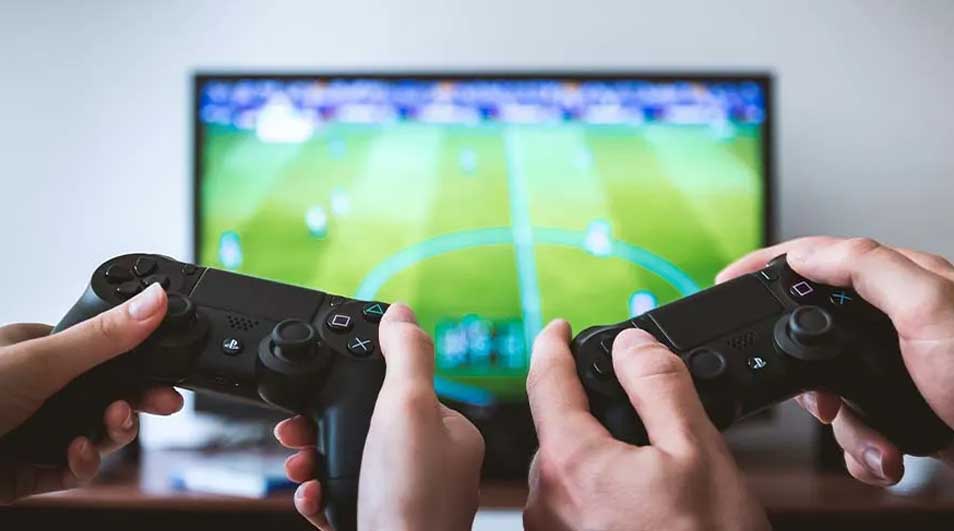 Twee handen houden zwarte gamecontrollers vast voor een tv-scherm met een groen voetbalveld.