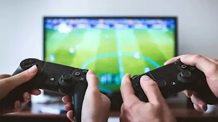 Twee handen houden zwarte gamecontrollers vast voor een tv-scherm met een groen voetbalveld.