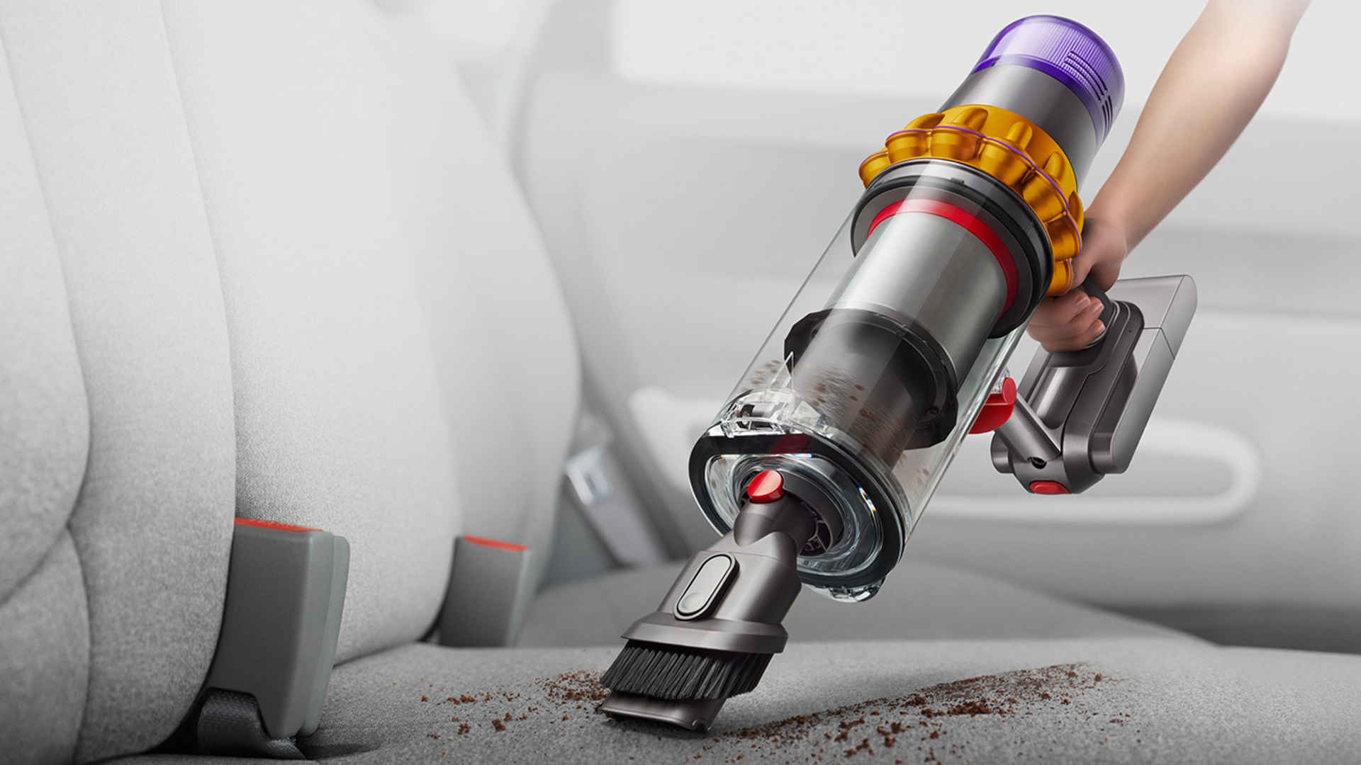 Een hand houdt een draadloze stofzuiger boven een grijze autostoel met kruimels. De stofzuiger heeft een paarse, gouden en rode accenten.