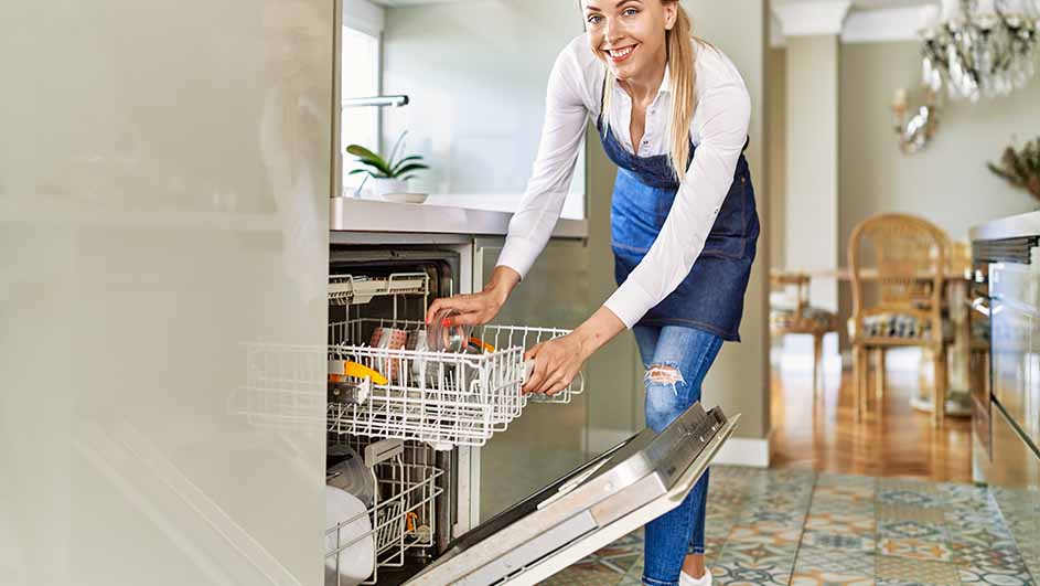 Een vrouw laadt de witte vaatwasser in een keuken met witte kasten en een tegelvloer.