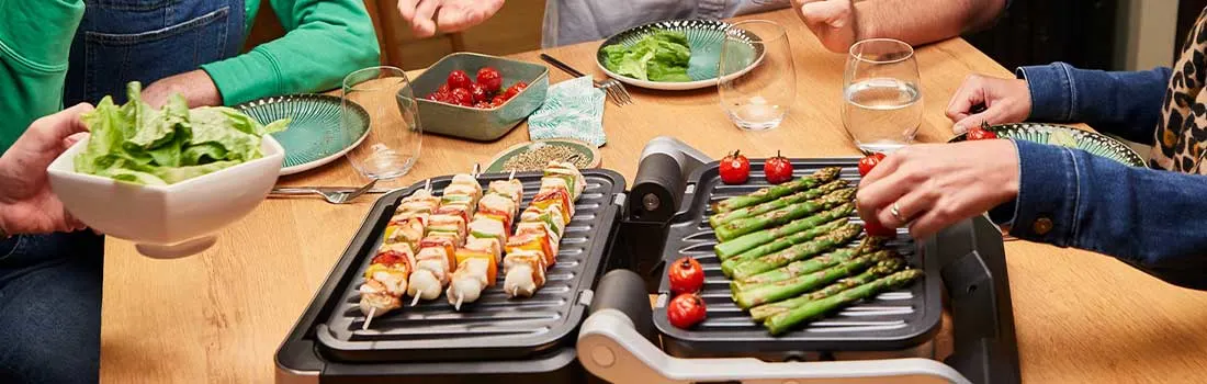 Aan een eettafel bereiden vrienden samen groentenspiesen en asperges op een tafelgrill, terwijl salades en drankjes gezellig worden gedeeld.