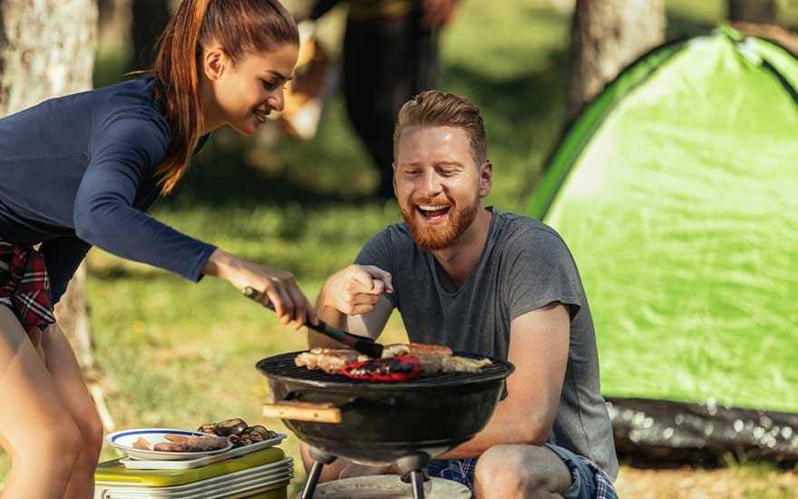 A cheerful couple barbecue at a campsite beside a green tent, as meat and vegetables sizzle on a small charcoal grill.