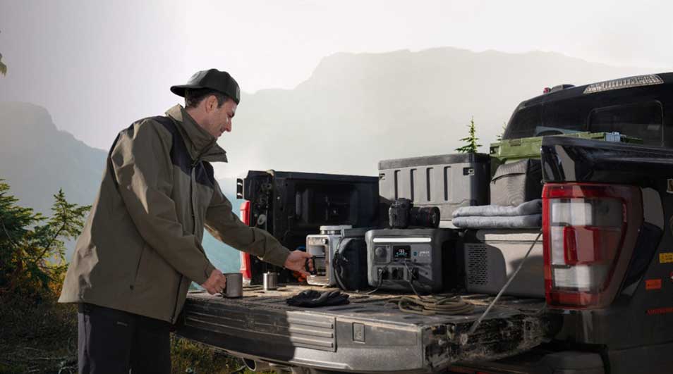 Een man bij een pick-up in de bergen gebruikt een draagbare stroomgenerator tussen kampeeruitrusting en koffers, omringd door natuur.