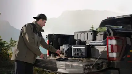 Een man bij een pick-up in de bergen gebruikt een draagbare stroomgenerator tussen kampeeruitrusting en koffers, omringd door natuur.