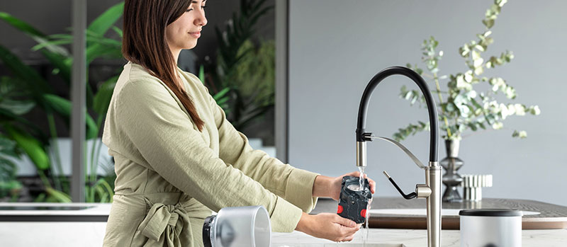 Woman rinsing a coffee machine part under a kitchen faucet in a modern kitchen.