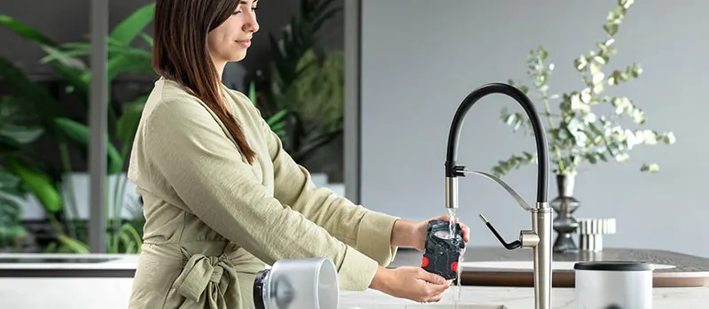 Woman rinsing a coffee machine part under a kitchen faucet in a modern kitchen.