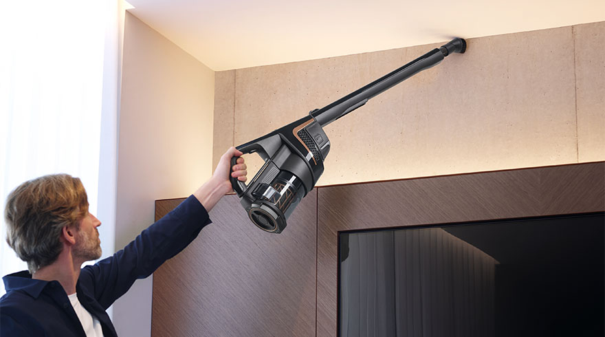 Een man gebruikt een draadloze stofzuiger met een lange buis tegen een muur boven een donker meubelstuk.