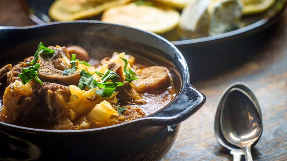 Een zwarte kom met stoofvlees, aardappelen en peterselie. Op de achtergrond een zwarte schaal met ronde, gele plakken. Twee lepels rechts.