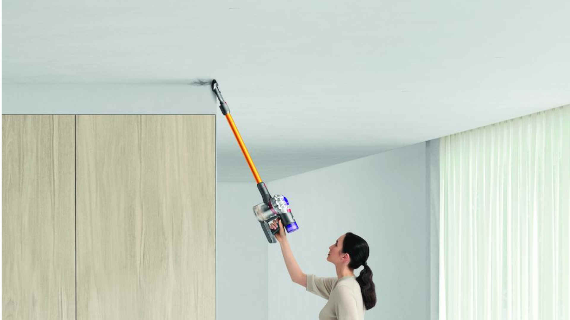 Een vrouw gebruikt een oranje stofzuiger om een wit plafond schoon te maken in een kamer met witte gordijnen en een kast.