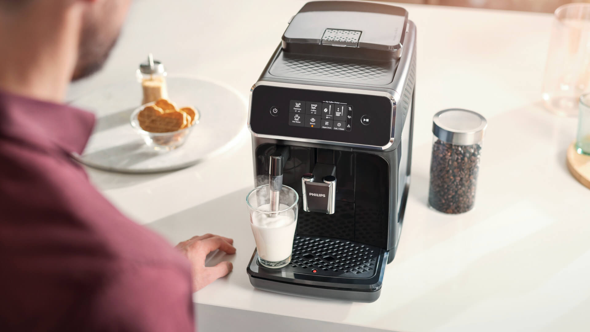Een zwarte Philips koffiemachine staat op een wit aanrecht, met een glas melk eronder. Een man staat ernaast.