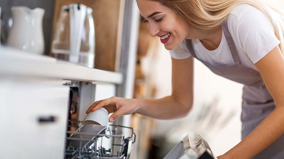 Vrouw laadt witte mok in vaatwasser in een keuken. Achter haar staat een witte kan en een glazen karaf.