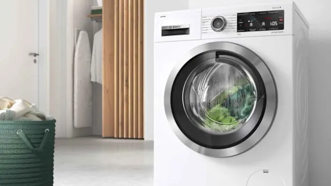 Witte wasmachine met groene was, metalen ring, digitaal display, groene wasmand links, houten wand en witte deur op achtergrond.