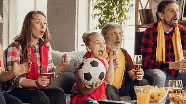 Een gezin juicht enthousiast in de woonkamer tijdens het kijken naar voetbal; een meisje houdt een bal vast, snacks en drank staan klaar.