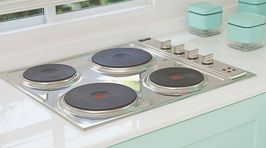 Een roestvrijstalen kookplaat met vier zwarte pitten staat op een lichtgroen aanrecht, naast blauwe dozen en een wit raam.