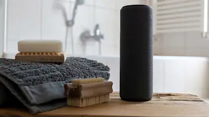 Een zwarte speaker, grijze handdoek, houten borstel en zeep op een houten oppervlak in een badkamer.