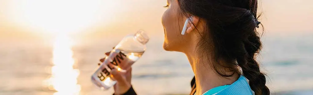 Vrouw met vlecht en draadloze oordopjes drinkt uit een fles water terwijl ze bij zonsondergang naar de zee kijkt. Op een warme avond buiten.