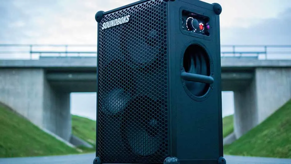 Zwarte rechthoekige luidspreker met rooster en bedieningselementen, buiten geplaatst met een brug op de achtergrond.