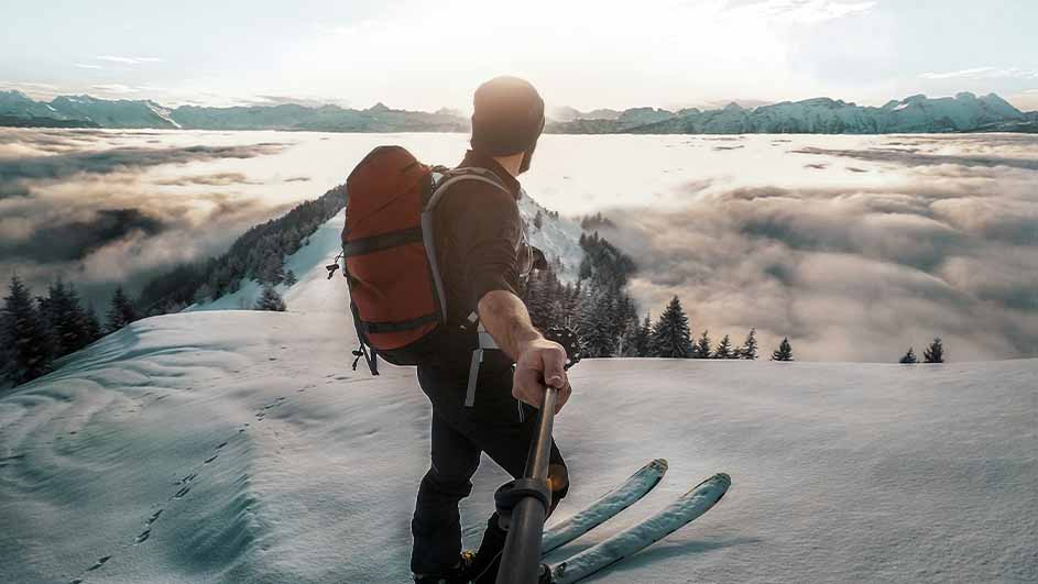 Een persoon met een rode rugzak staat op ski's in de sneeuw met bergen en wolken op de achtergrond.